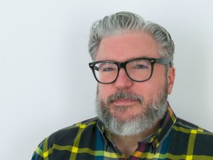 Photo of author Alonso Duralde. The author has salt and pepper hair, mustache, and beard, and wears black glasses. He is looking at the camera in a candid shot.